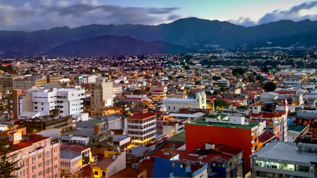 Aerial view of a brightly lit cityscape with colorful buildings at dusk, backed by a range of mountains under a cloudy sky.